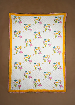 A rectangle quilted textile with an off-white center printed with colorful hot air balloons and clouds, finished with a wide mustard yellow border, laid flat against a brown background.