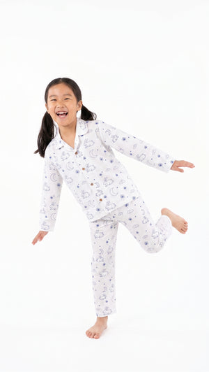 A young girl with dark hair tied back is wearing a white long-sleeve pajama set with a blue cartoon cloud pattern, captured in a playful pose against a solid white background.