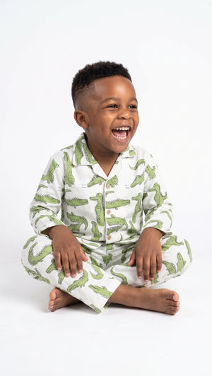 A young child sitting cross-legged on a white studio background, wearing a pajama set with a green crocodile print on a white base.