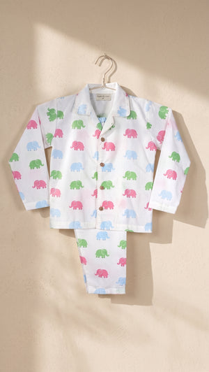 A children's long-sleeve pajama set with matching pants, featuring a white base printed with small colorful elephant silhouettes in pink, green, blue, and light blue, hanging on a hanger against a warm beige wall with soft sunlight and shadows.