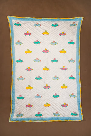A rectangle quilted baby quilt with a white background, featuring a repeating pattern of colorful cartoon cars and a light blue border with a yellow twisted rope trim, displayed flat against a brown background.