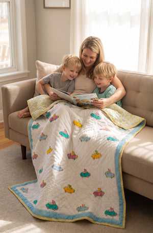 A woman and two young boys sit on a beige sofa in a bright living room, reading a book while covered under a white quilted blanket with a light blue border and colorful car-print motifs.