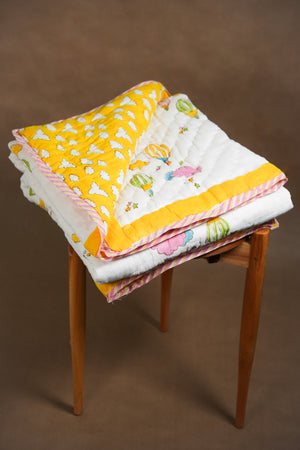 A folded quilted baby blanket with a yellow and white cloud print and a white side with colorful hot air balloon motifs, displayed on a wooden stool against a brown background.