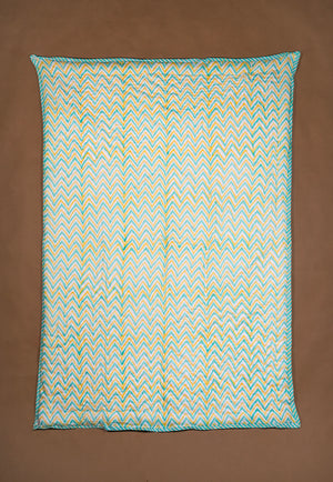 A rectangular quilted blanket with a colorful pattern in teal, yellow, and white, displayed flat against a solid brown background.