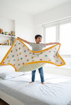 A child stands on a white bed in a bright bedroom, holding up a large quilted blanket with a yellow border and colorful tree pattern.