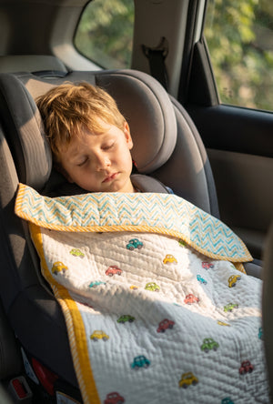 A young child sleeping peacefully in a gray car seat, wrapped in a white quilted blanket with colorful car patterns and yellow trim.