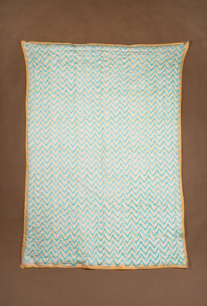 A rectangle quilt, featuring a light blue and white pattern with a yellow-orange braided border, laid flat against a solid brown background.