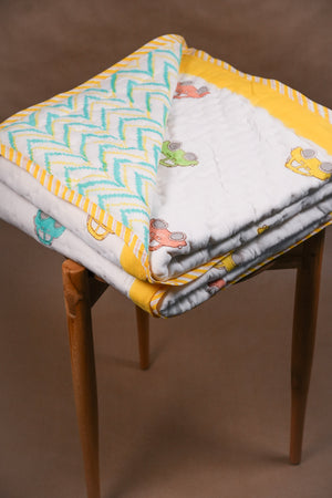 A folded quilted baby blanket with a white base, yellow chevron trim, and colorful car prints, displayed on a wooden stool against a brown background.