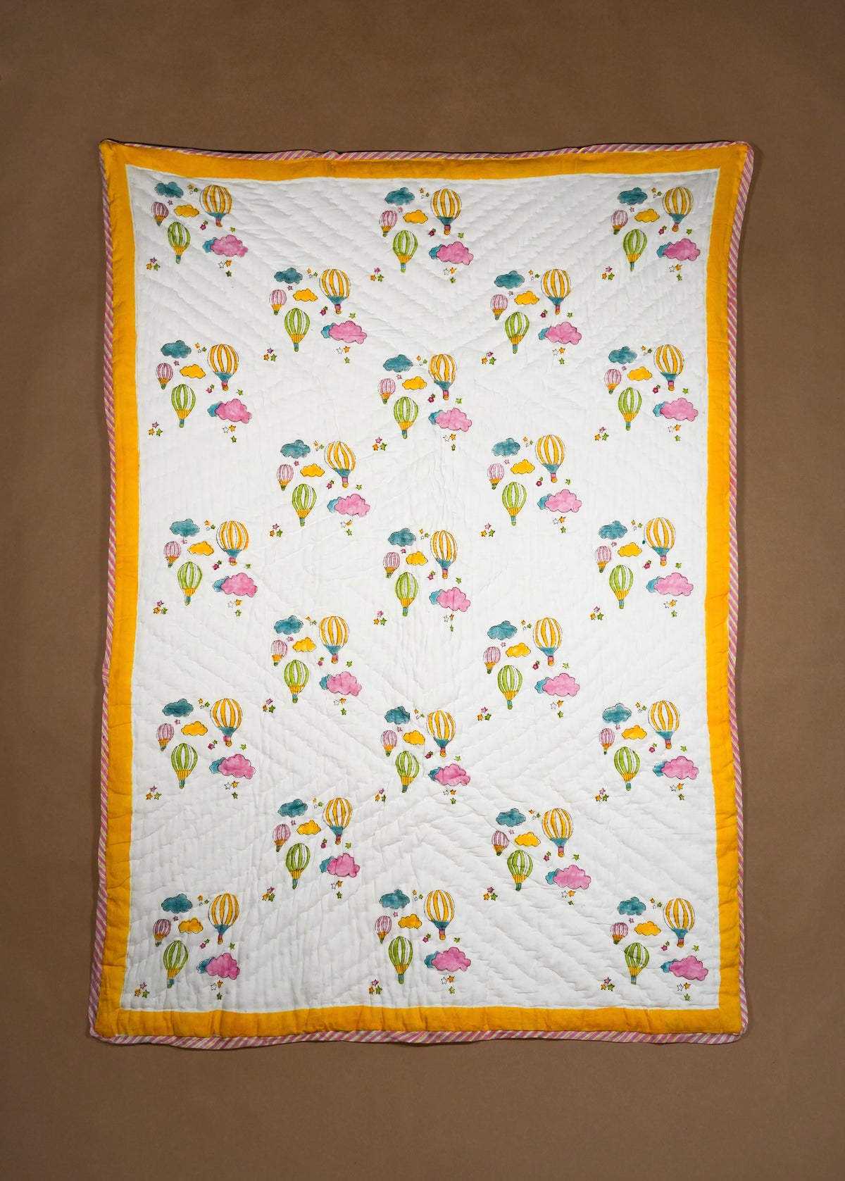 A rectangle quilted textile with an off-white center printed with colorful hot air balloons and clouds, finished with a wide mustard yellow border, laid flat against a brown background.