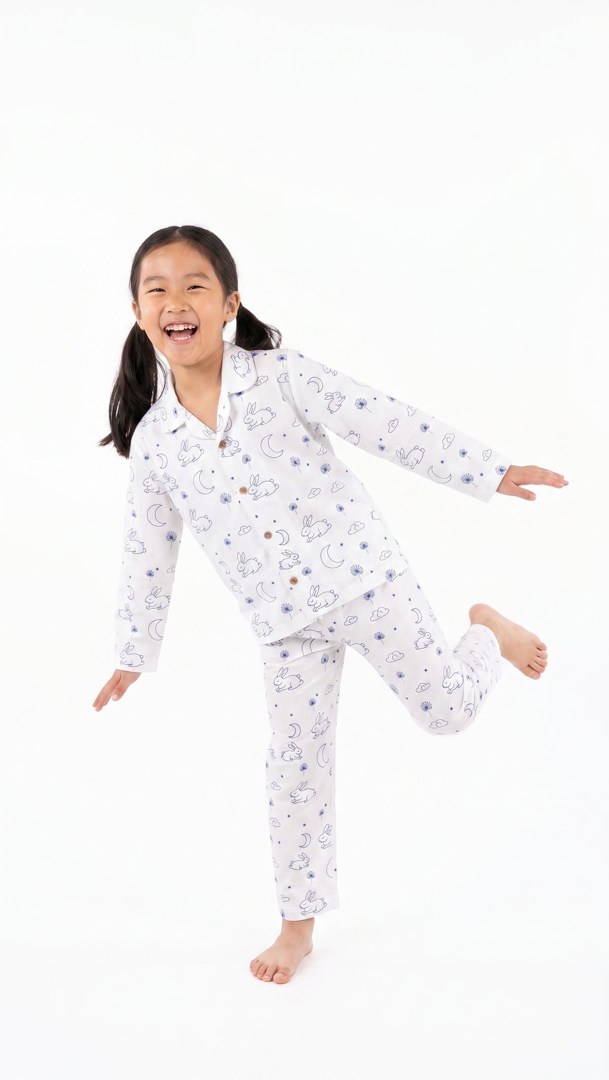 A young girl with dark hair tied back is wearing a white long-sleeve pajama set with a blue cartoon cloud pattern, captured in a playful pose against a solid white background.