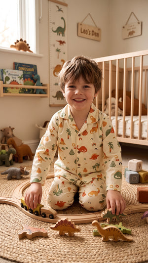 A smiling young boy sits on a woven rug in a child's bedroom, wearing cream pajamas with a colorful dinosaur print. He is surrounded by wooden toy train tracks and dinosaur blocks.
