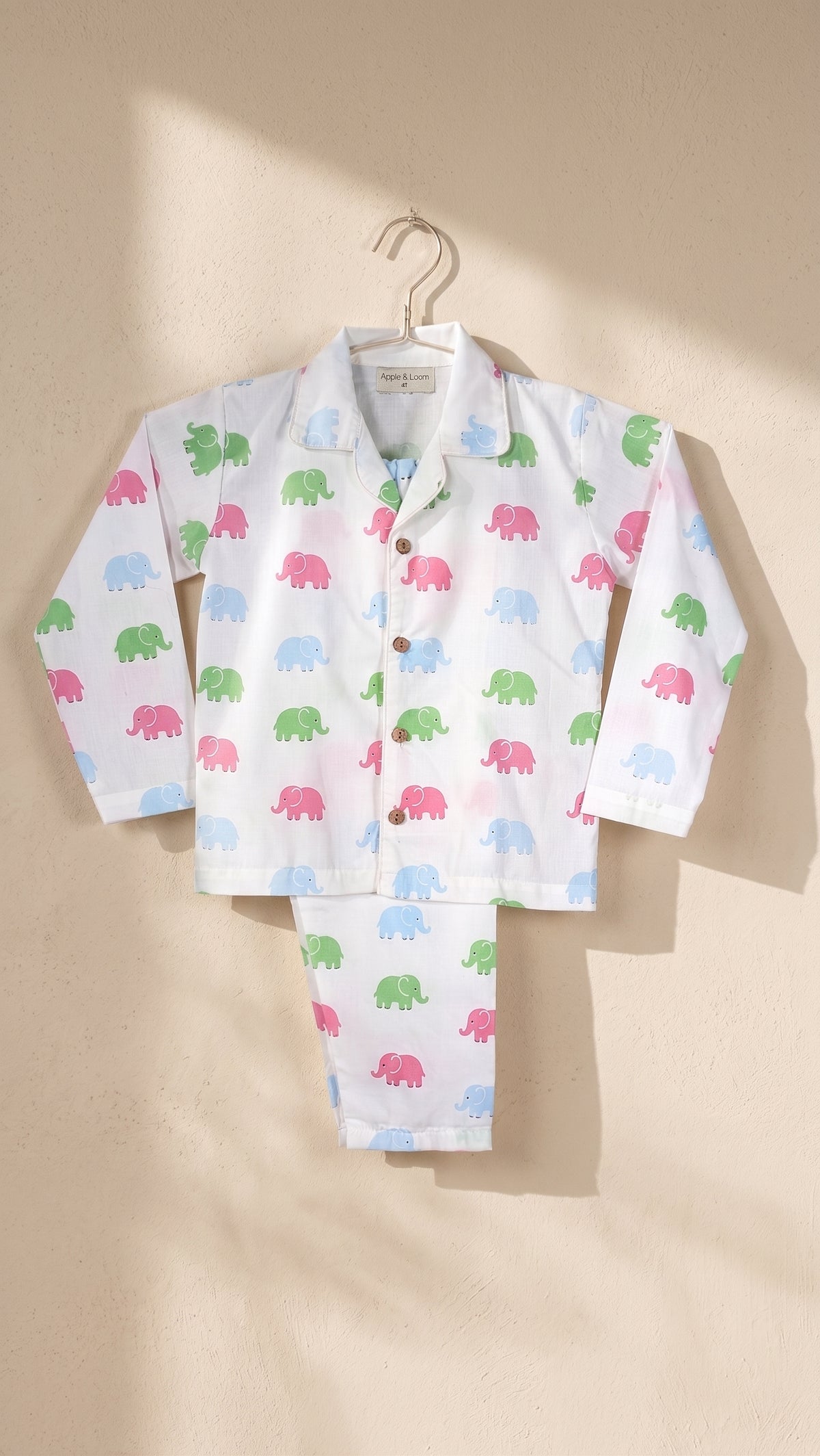 A children's long-sleeve pajama set with matching pants, featuring a white base printed with small colorful elephant silhouettes in pink, green, blue, and light blue, hanging on a hanger against a warm beige wall with soft sunlight and shadows.