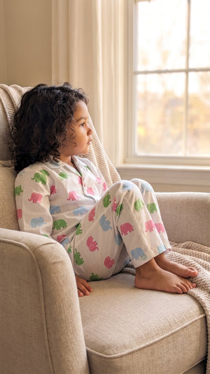 A child sits barefoot on a beige armchair by a window, wearing white pajamas with a colorful elephant print.