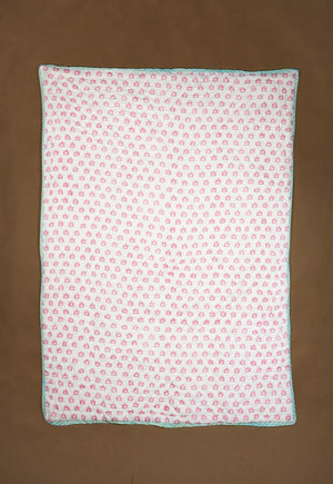 A rectangle quilted baby blanket with a pink and white floral pattern and a light blue border, laid flat on a brown background.