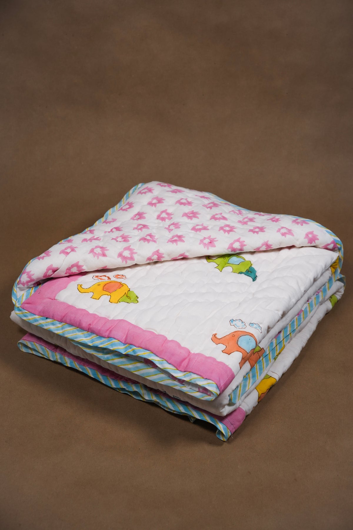 A folded quilted baby blanket with a white background, pink floral pattern on one side, colorful cartoon elephants on the other, and pink and blue striped borders, set against a solid brown background.