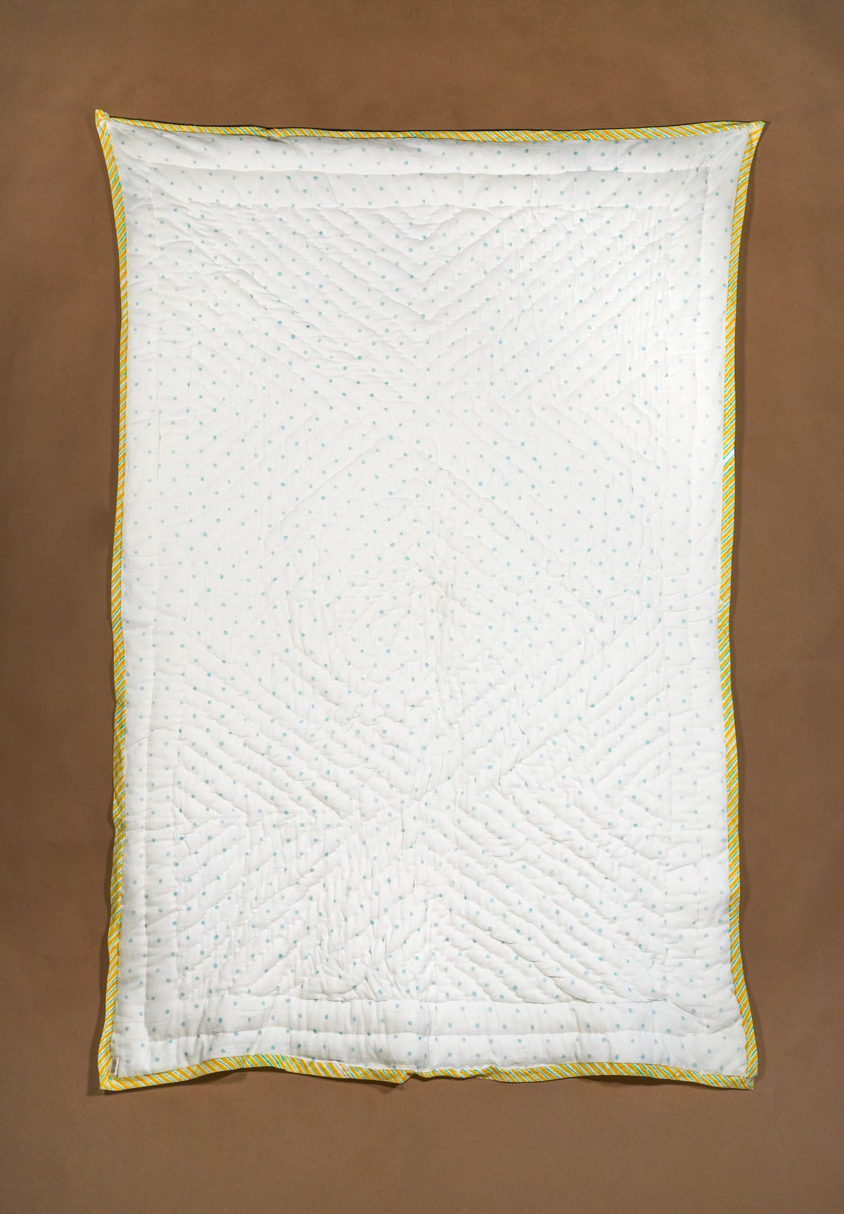 A white quilted baby quilt with blue dots and a yellow and white twisted rope border, laid flat on a brown background.