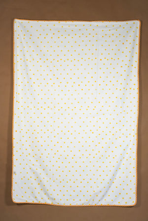 A square white fabric item, likely a blanket or towel, featuring a small yellow polka dot pattern and a yellow braided border, laid flat against a solid brown background.
