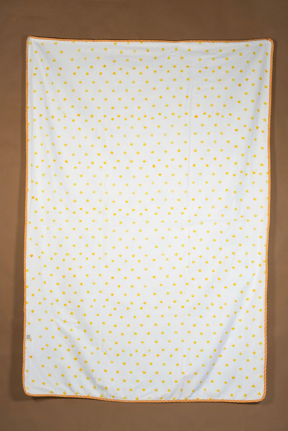 A square white fabric item, likely a blanket or towel, featuring a small yellow polka dot pattern and a yellow braided border, laid flat against a solid brown background.