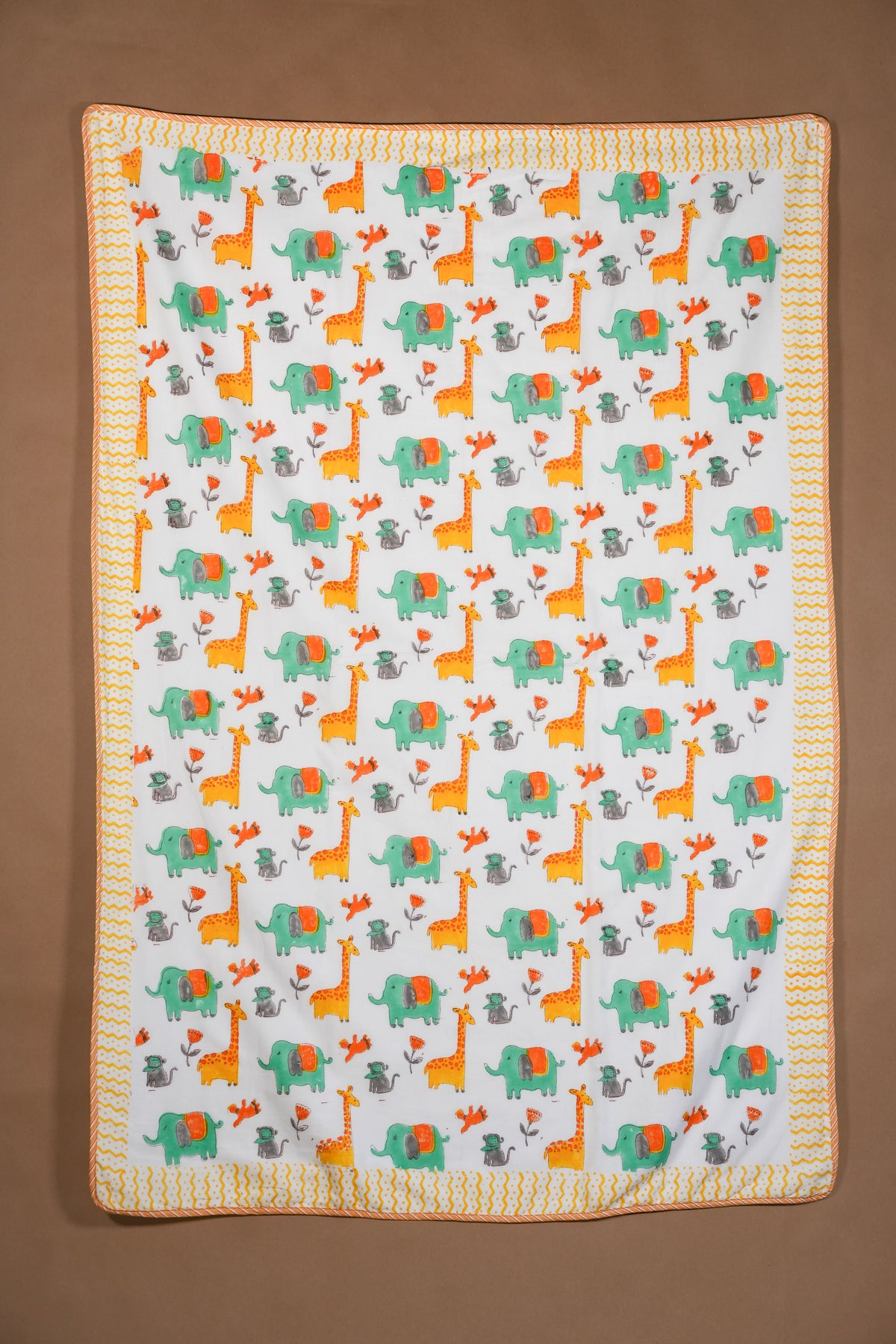 A square baby blanket with a white background featuring a repeating pattern of orange giraffes, green elephants, and gray acacia trees, finished with a yellow and orange zigzag border, shown flat against a brown background.