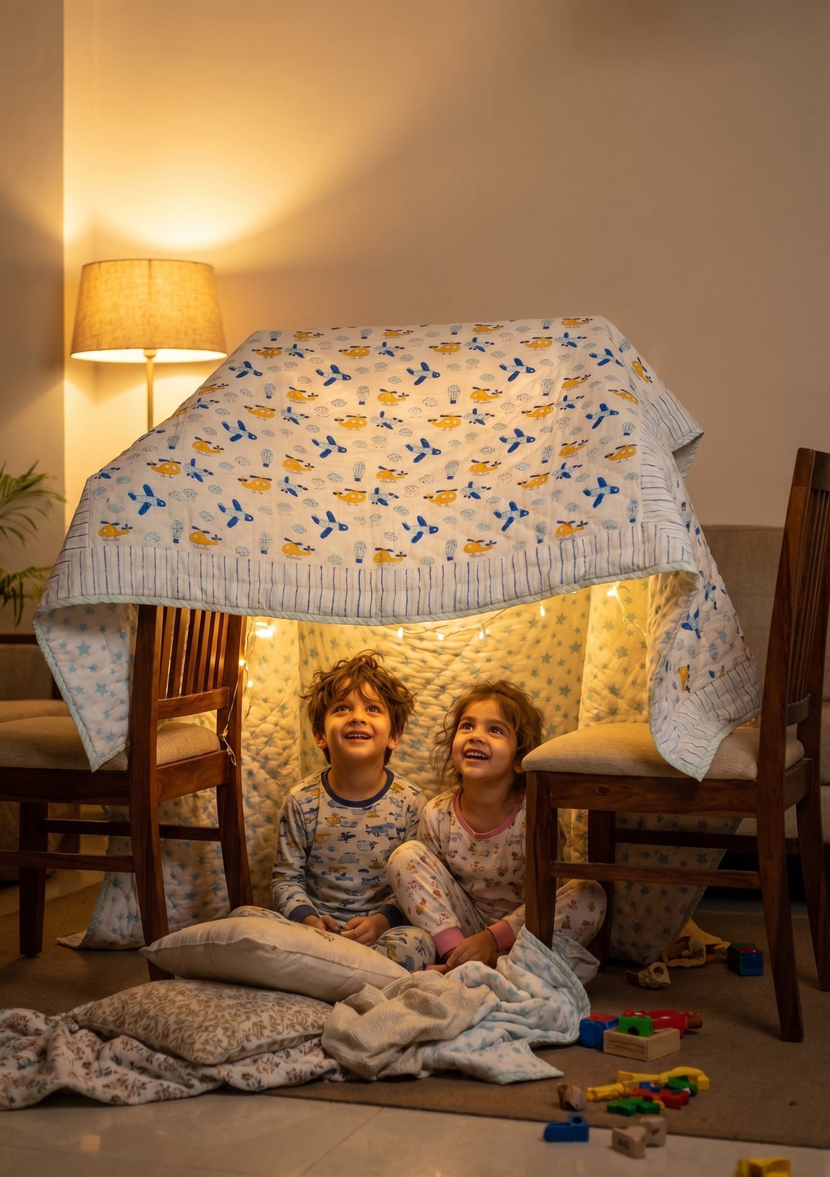 Two children sitting under a quilted play tent with string lights in a warmly lit room, with chairs and toys visible around them.