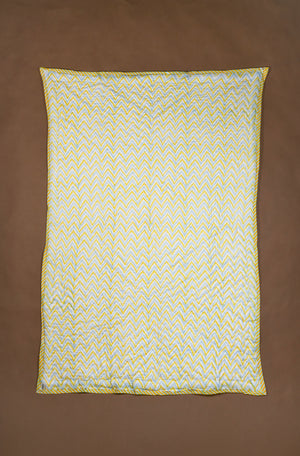 A rectangle quilted blanket with a yellow and white chevron pattern and a yellow border, laid flat on a solid brown background.