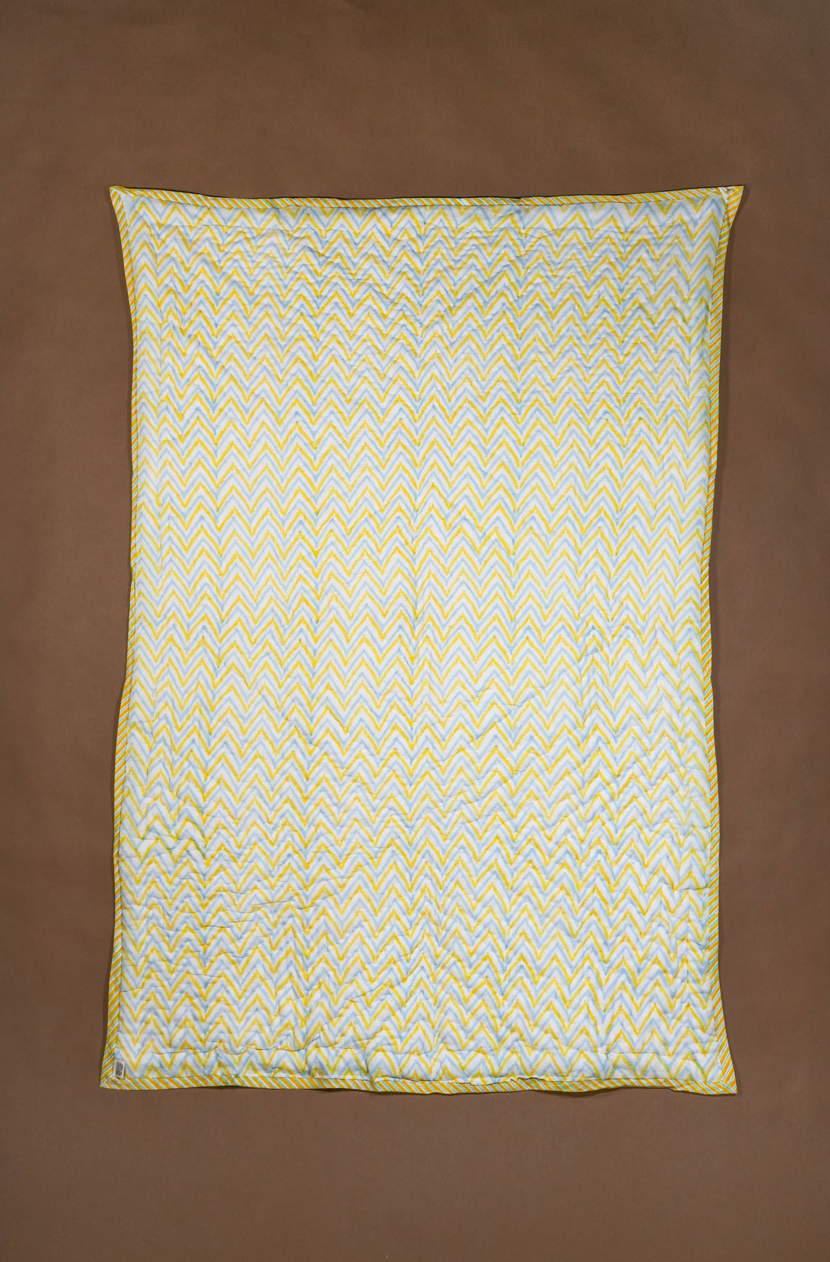 A rectangle quilted blanket with a yellow and white chevron pattern and a yellow border, laid flat on a solid brown background.
