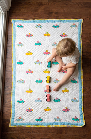 A baby sits on a quilted white play mat with a blue border and colorful car pattern, playing with wooden toy cars on a dark wood floor.