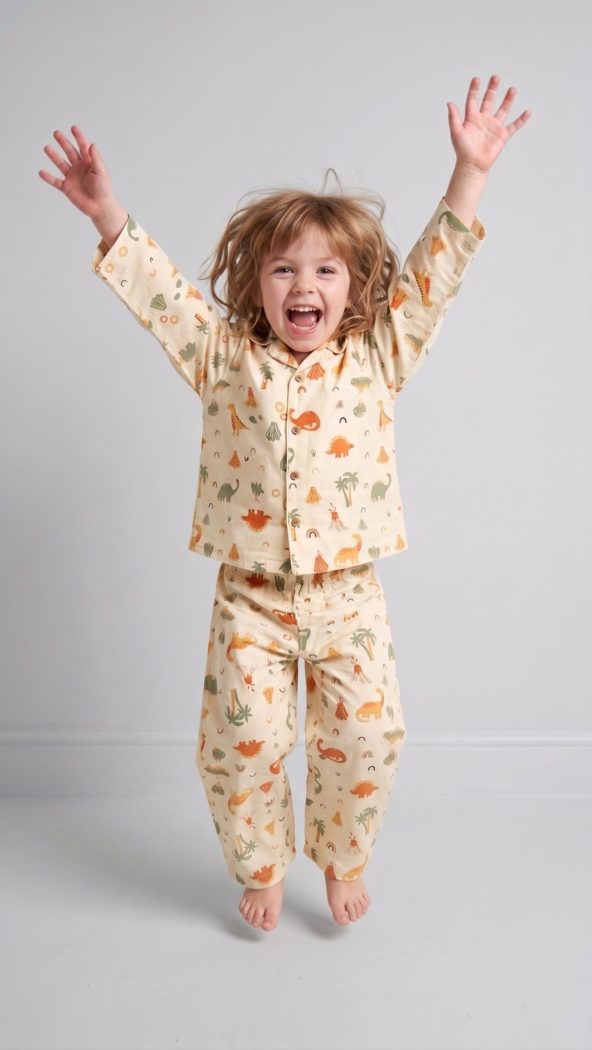 A young child with light brown hair jumps barefoot against a light gray studio background, wearing a cream-colored pajama set with a dinosaur pattern.