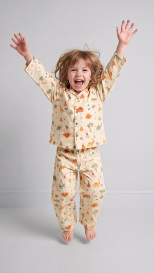 A young child with light brown hair jumps barefoot against a light gray studio background, wearing a cream-colored pajama set with a dinosaur pattern.