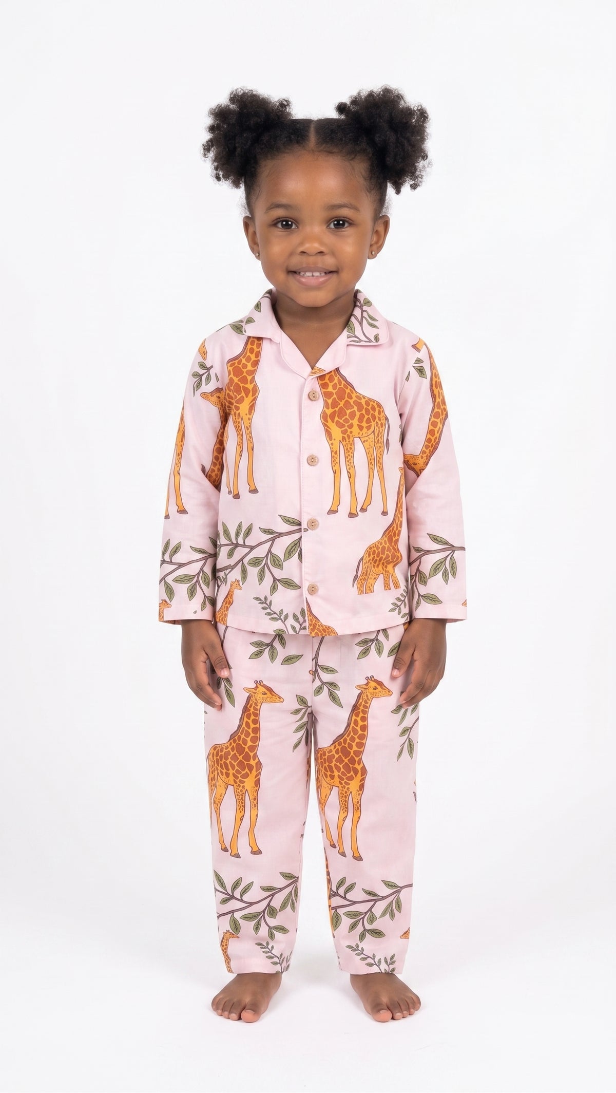 A young child wearing a two-piece pajama set with an orange giraffe and green leaf pattern on a light pink background, standing against a white studio background.