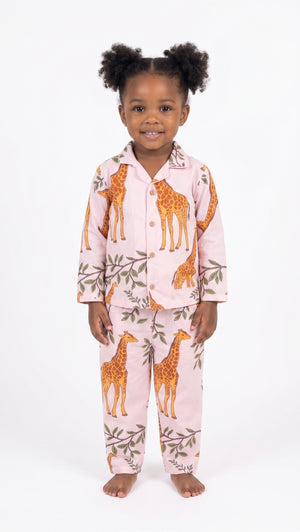 A young child wearing a two-piece pajama set with an orange giraffe and green leaf pattern on a light pink background, standing against a white studio background.