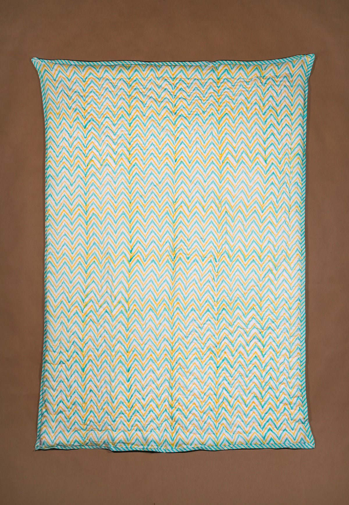 A rectangular quilted blanket with a colorful pattern in teal, yellow, and white, displayed flat against a solid brown background.