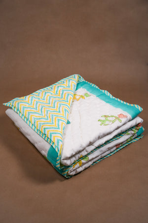 A folded quilted baby blanket with a white center, yellow and teal pattern on one side, and embroidered cartoon animals on a teal border, set against a solid brown background.