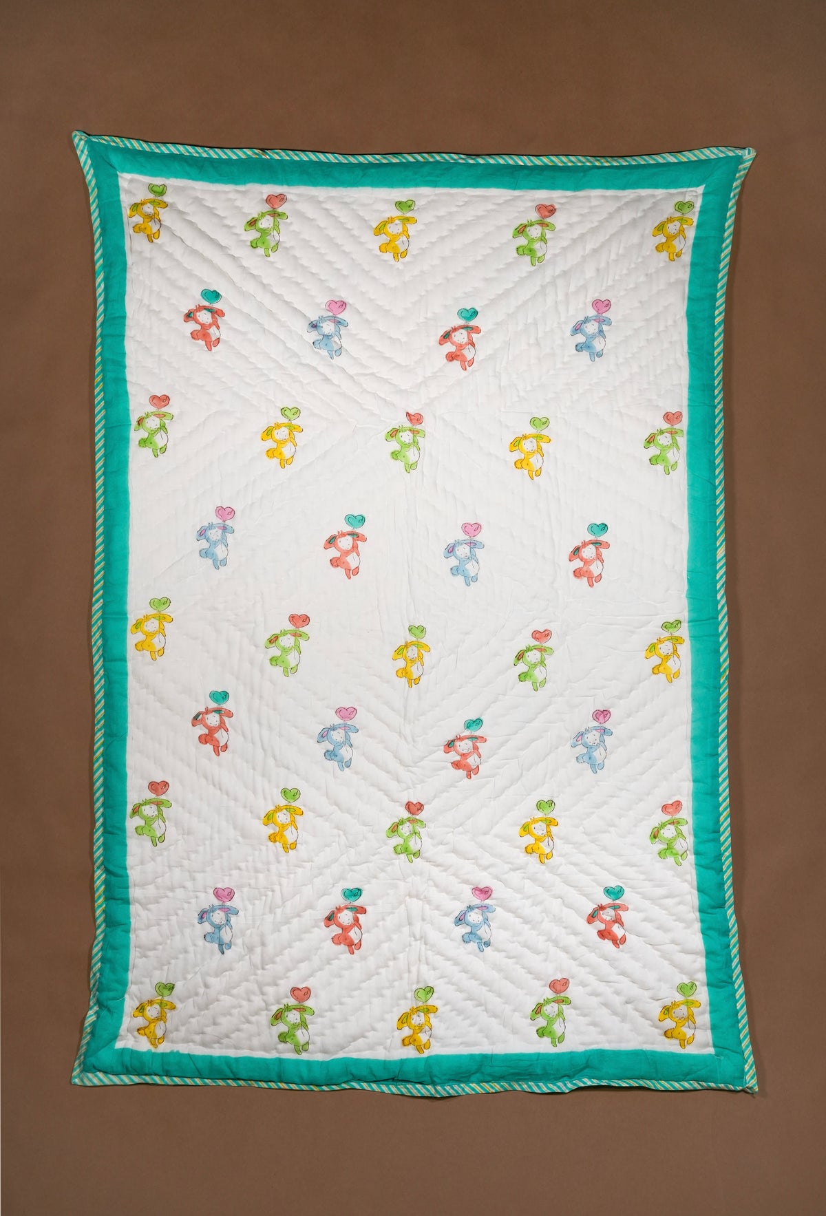 A rectangle white quilted blanket featuring a repeating pattern of colorful dancing figures and a bright teal border, displayed flat against a solid brown background.