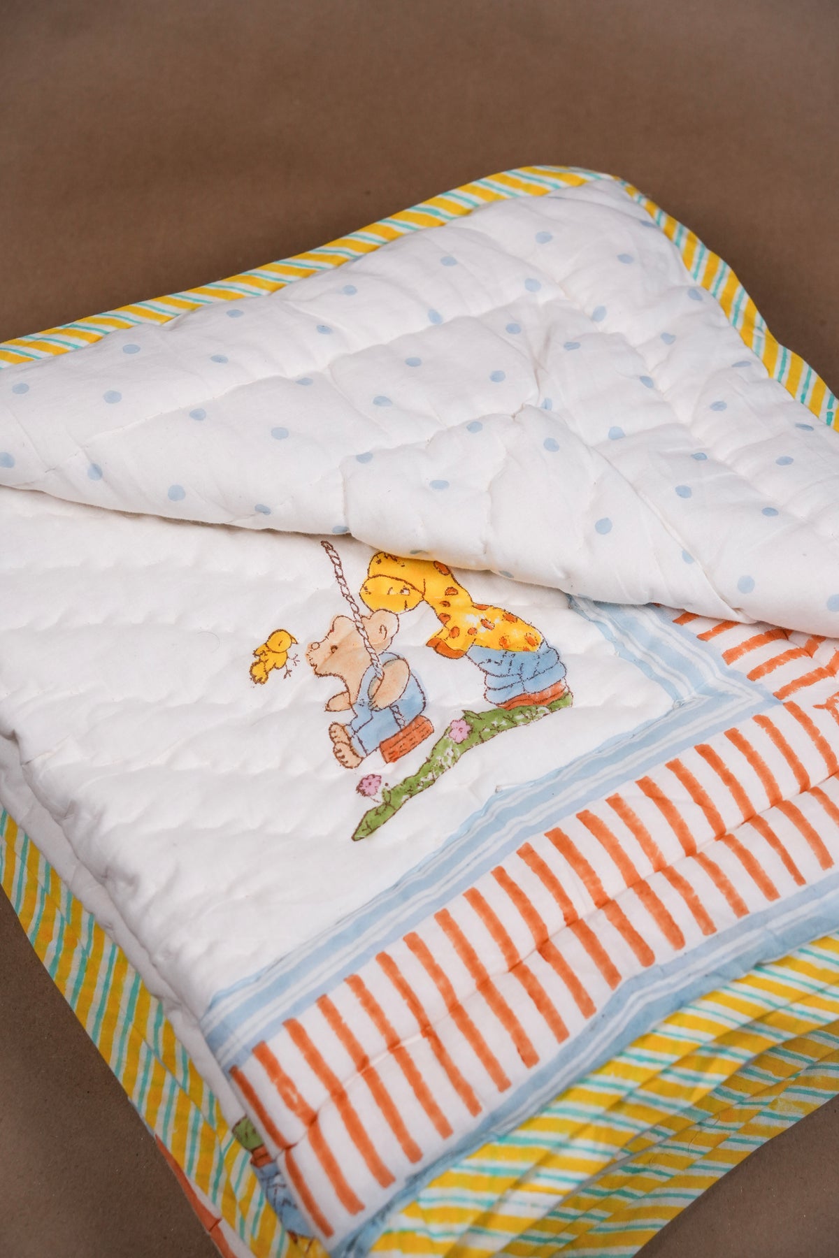 A folded quilted baby blanket with a white base, light blue polka dots, orange and white stripes, and a colorful embroidered cartoon bear motif, shown on a brown background.