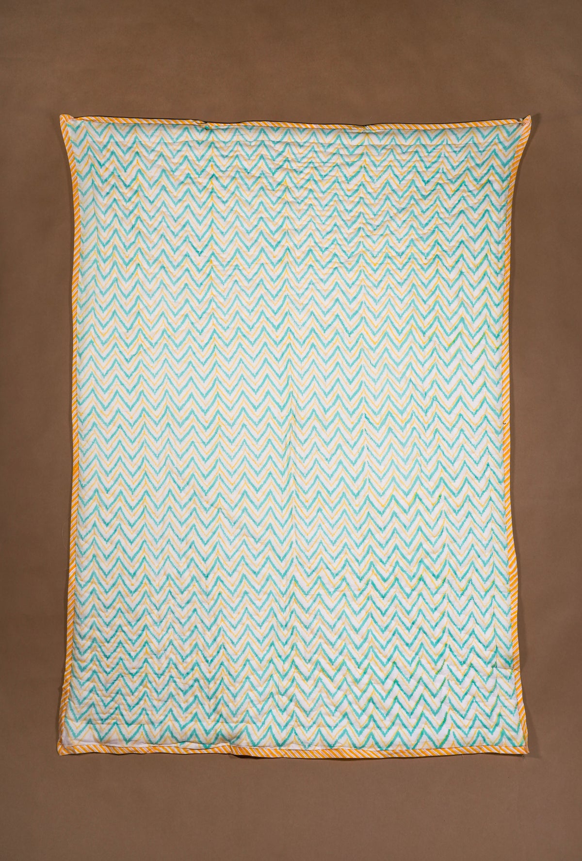 A rectangle quilt, featuring a light blue and white pattern with a yellow-orange braided border, laid flat against a solid brown background.