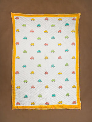 A baby quilted blanket with a white center, colorful embroidered cars, and a bright yellow border, laid flat on a brown background.